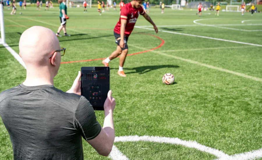 Dr Devon Lewis standing by a football pitch as a player kicks the ball. Devon is holding an iPad that shows live data streamed from a wearable sensor.