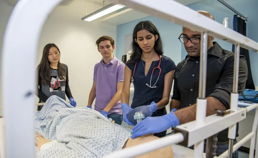 A group of students watch as one of them works on a dummy patient in a hospital bed