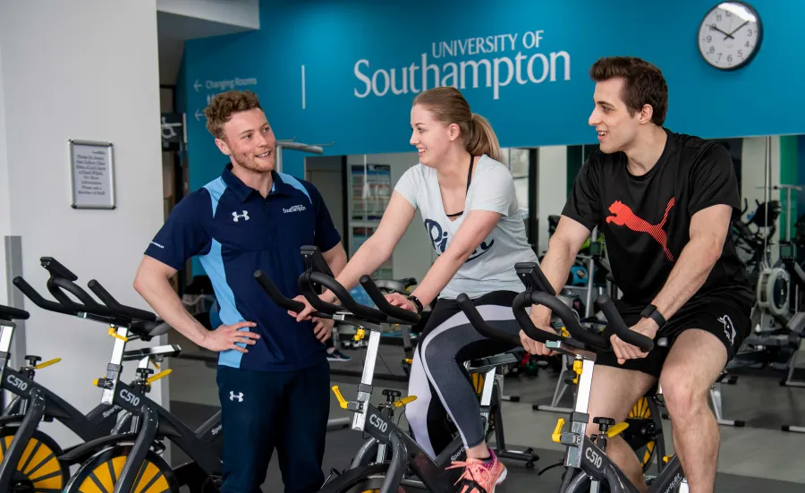 Two students working out on exercise bikes in a gym, assisted by a personal trainer.