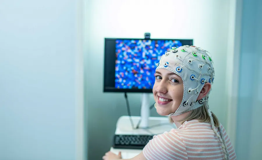 A student sits in front of a computer and turns to smile towards the camera. She is wearing a head cap that is connected to electrodes to monitor brain activity.