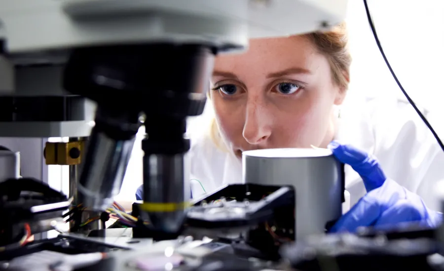 A female researcher peering intently at the contents of a slide in a high-powered microscope.