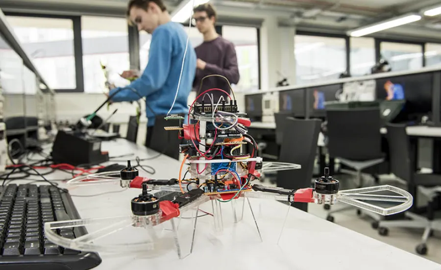 A close-up of a small, 4-rotor drone on a work bench in the laboratory. Two students in the background are testing electronics.