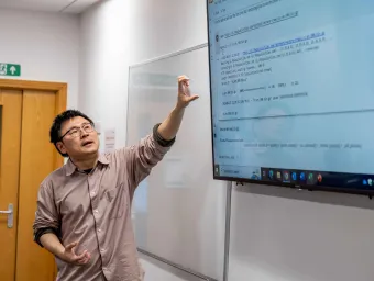 A lecturer extends their arm upwards, towards notes on a large screen mounted on the wall.