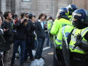 A crowd of protestors to the left, and a group of riot police to the right, confront each other. In the foreground, a punk in a leather jacket looks rebelliously across at one police officer who is raising their baton 
