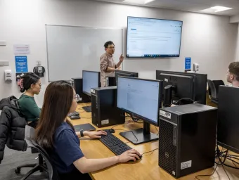 Three students in front of monitors in a classroom listen as a lecturer makes a point