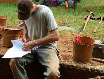An archaeologist sits on the edge of a freshly dug trench, making notes on a clipboard
