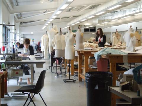 A wide shot of the fashion studio, full of students hard at work. To the right, a row of large workbenches are mixed in with over a dozen mannequins wearing various designs. To the left, a row of sewing machines.
