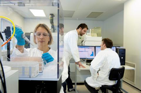 Researcher pipetting in a lab while two researchers talk in the background