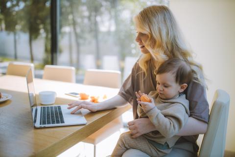 A woman sitting at a table using a laptop with one hand and a child sitting on her knee eating an orange 