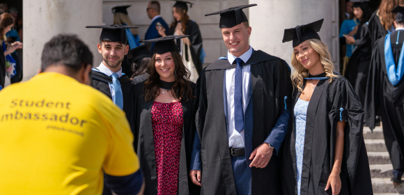 A student ambassador in a yellow tshirt takes a photograph of four graduands posing in front of the O2 Guildhall.