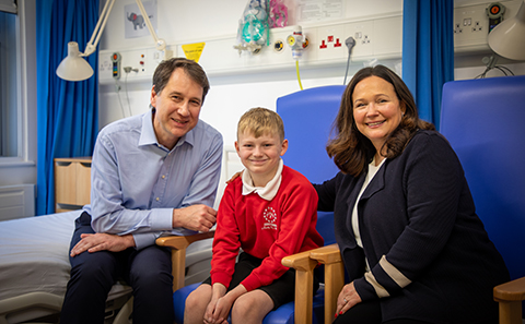  Boy in red jumper sat between a man and a woman in a hospital ward.