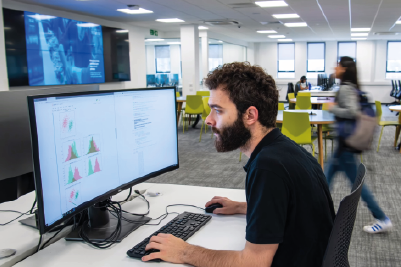 A student using a computer in a shared workspace. On his screen are a set of graphs in pink and green. Another student is walking past in the background.
