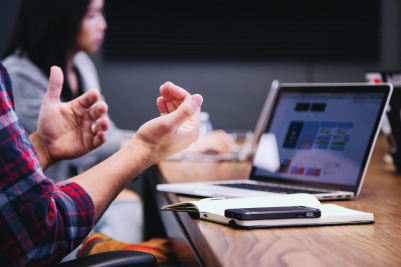 A student gesturing with their hands in front of their laptop with another student in the background.
