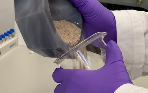 Close up of someone wearing a lab coat and blue latex gloves pouring grey-coloured powder from a grey bowl into a clear glass container