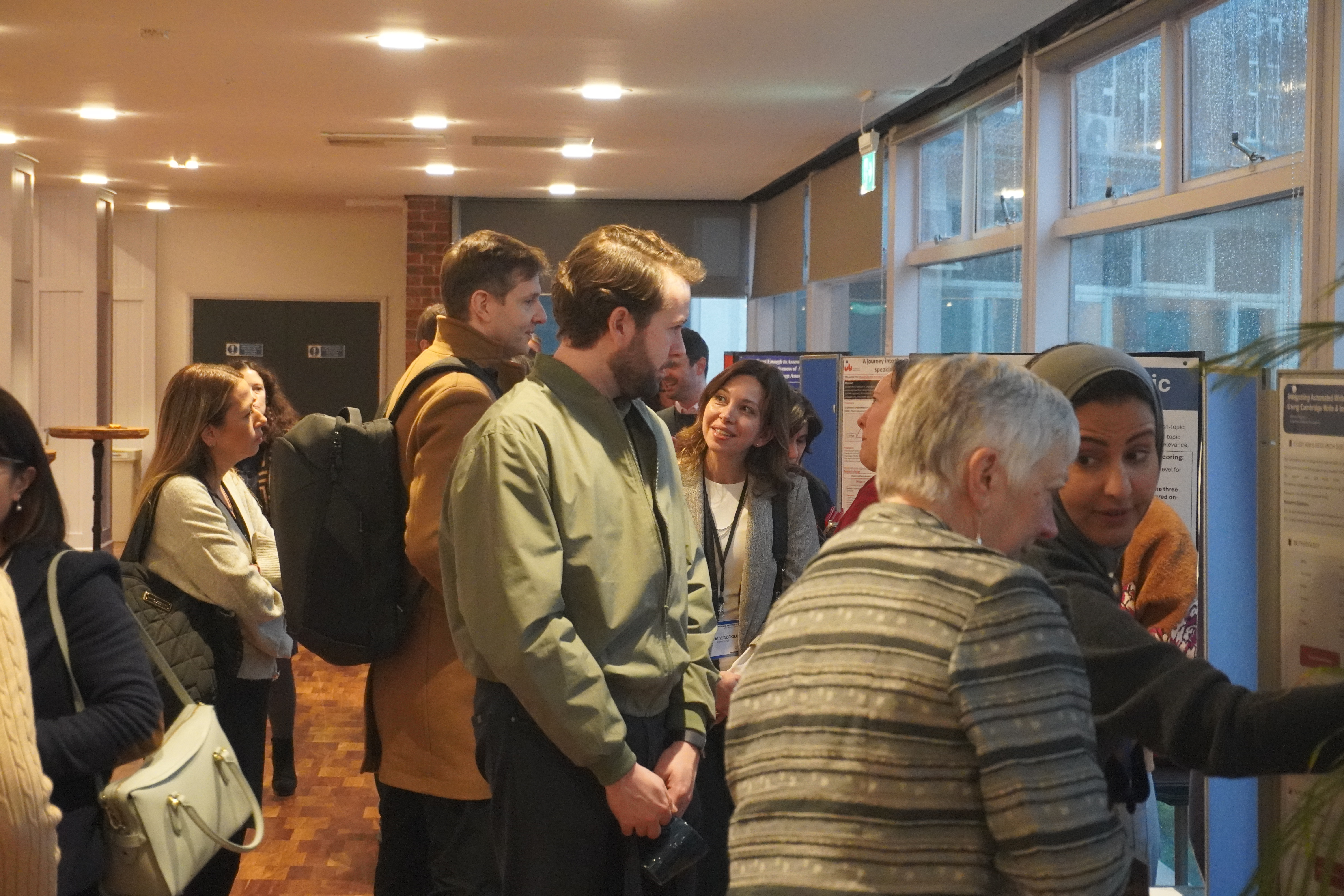 Several people are gathered in a hallway, viewing and discussing posters displayed on boards near large windows.