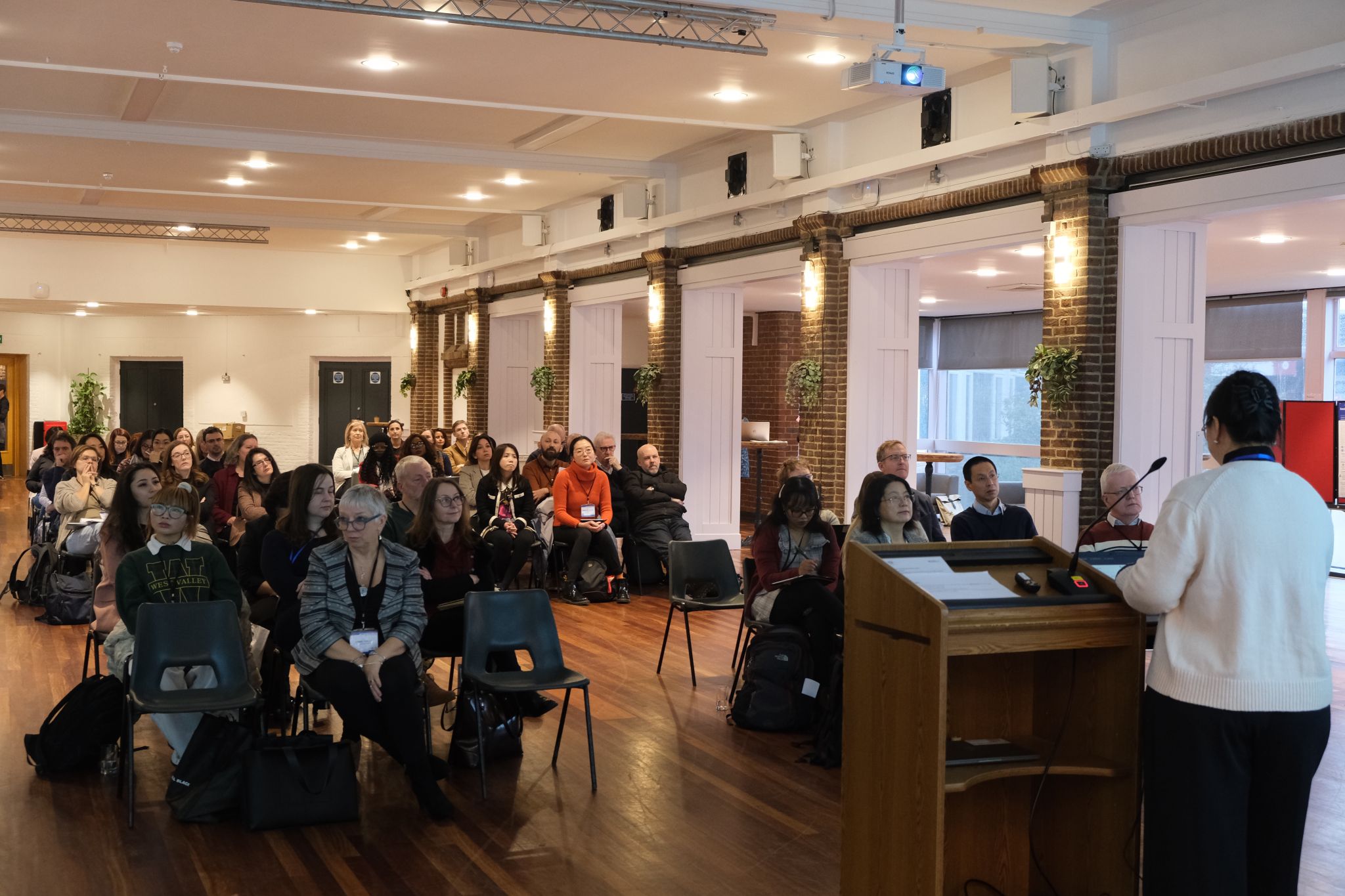 A speaker addresses an audience seated in rows inside a bright conference room with wooden floors and brick accents.