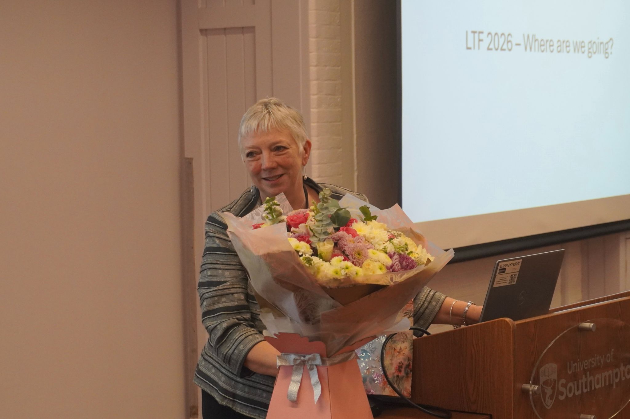 A person stands at a podium holding a large bouquet of flowers, with a presentation slide in the background that reads “LTF 2026 – Where are we going?”