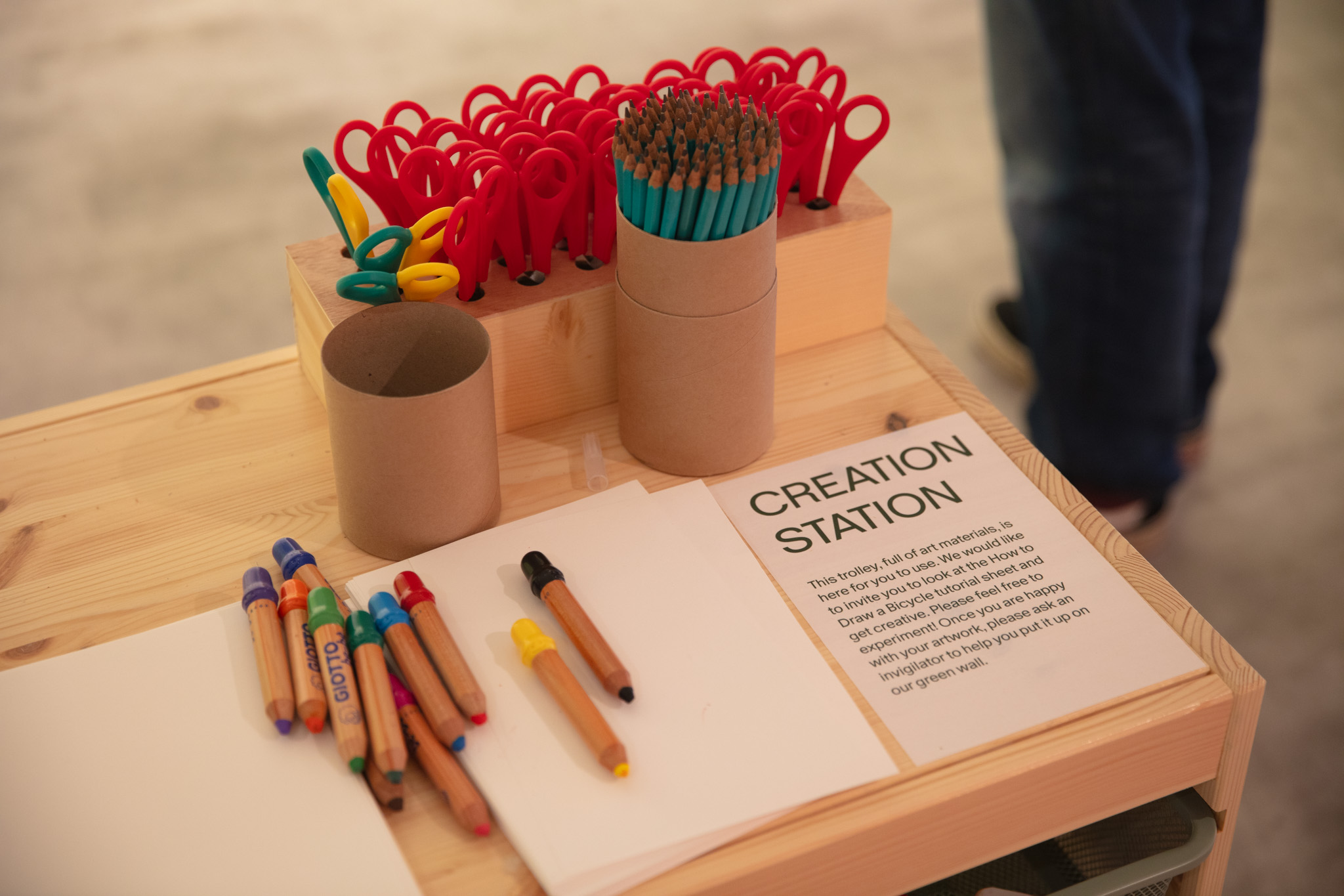 Plain paper, pots of scissors and pens and pencils sit on a table which is labelled as a 'creation station'