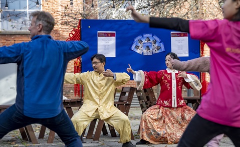 Man and woman in traditional Chinese dress performing tai chi, with members of the public joining in.