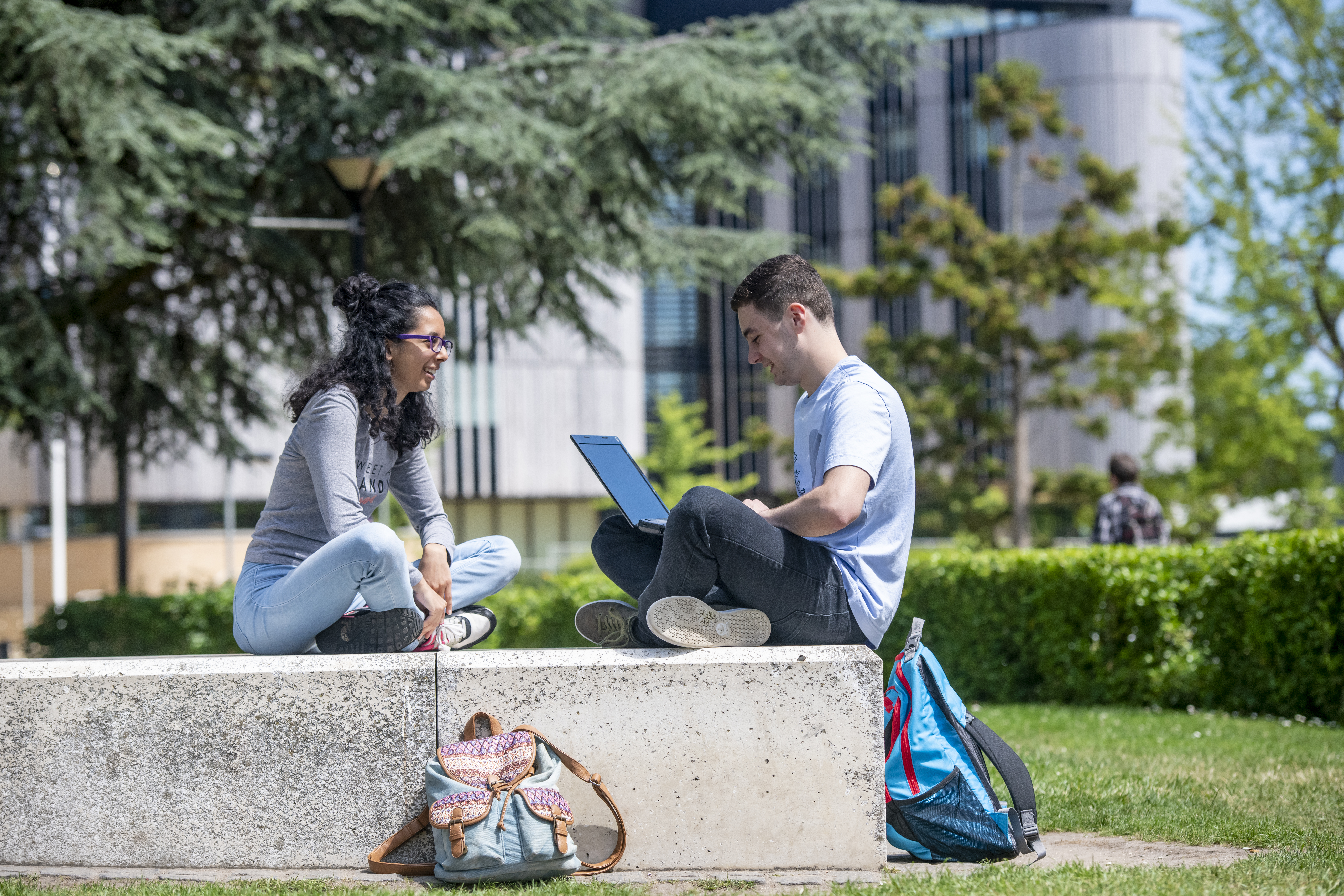 Two students sit outdoors on a concrete bench on a university campus, talking while one uses a laptop. Backpacks rest nearby, with trees, green lawns and a modern academic building in the background.