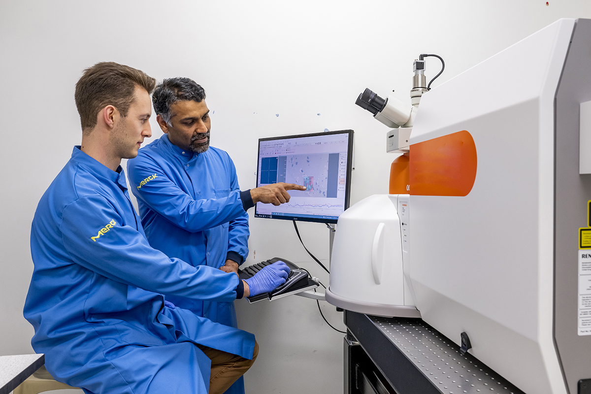 Researchers working on a rapid laser-based test that could diagnose different types of dementia in minutes. Two scientists in blue lab coats are working in a laboratory. One is seated, wearing gloves, typing on a keyboard, while the other is standing, pointing at a computer monitor displaying scientific data. They are using a large white and orange analytical instrument, likely a spectrometer or microscope, for research or analysis.