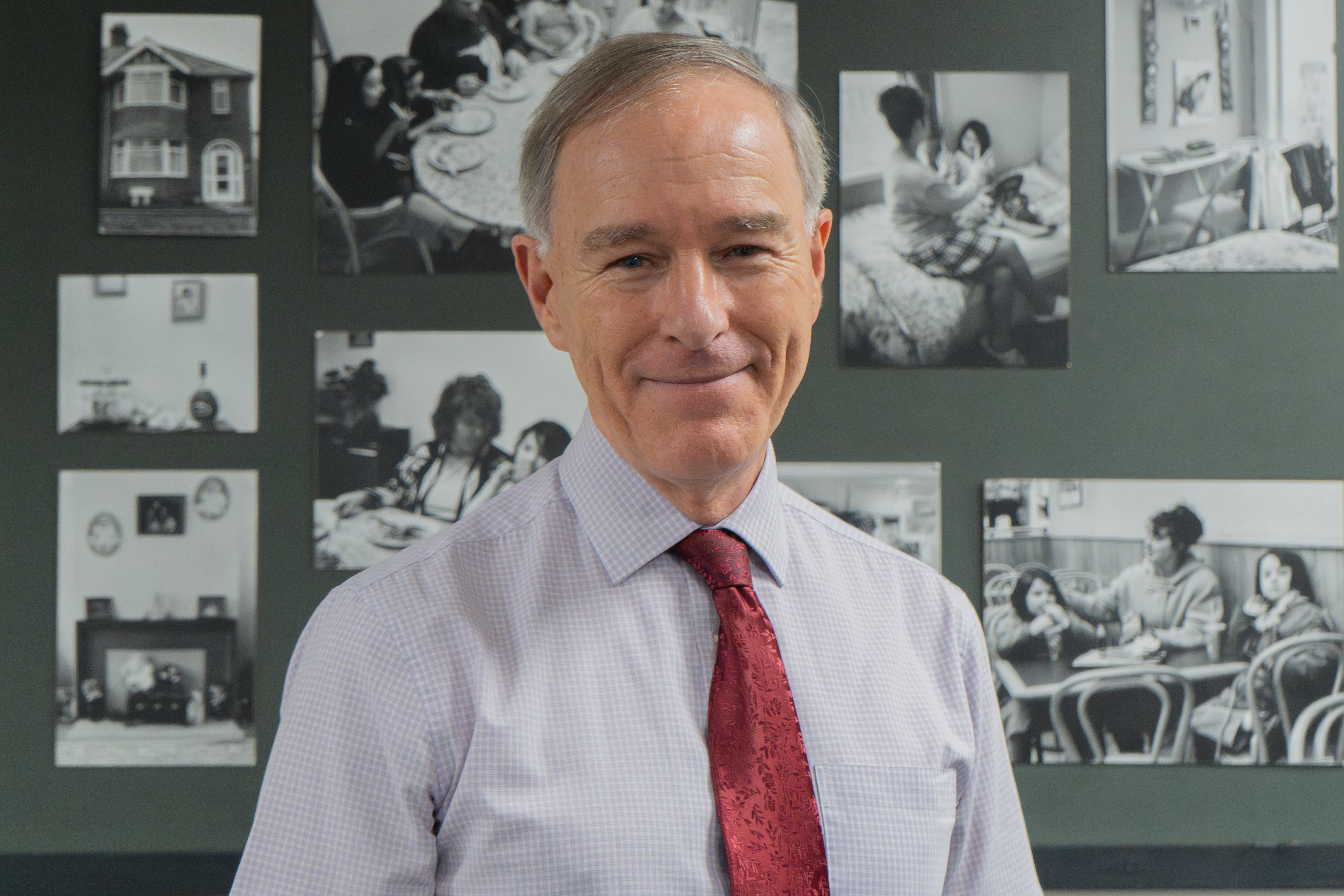 A middle-aged man wearing a light-coloured shirt and red tie stands indoors in front of a wall displaying several black-and-white photographs. He is facing the camera and smiling slightly.
