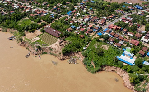 Aerial view of a village beside a muddy river, with houses, greenery, and boats on the riverbank.