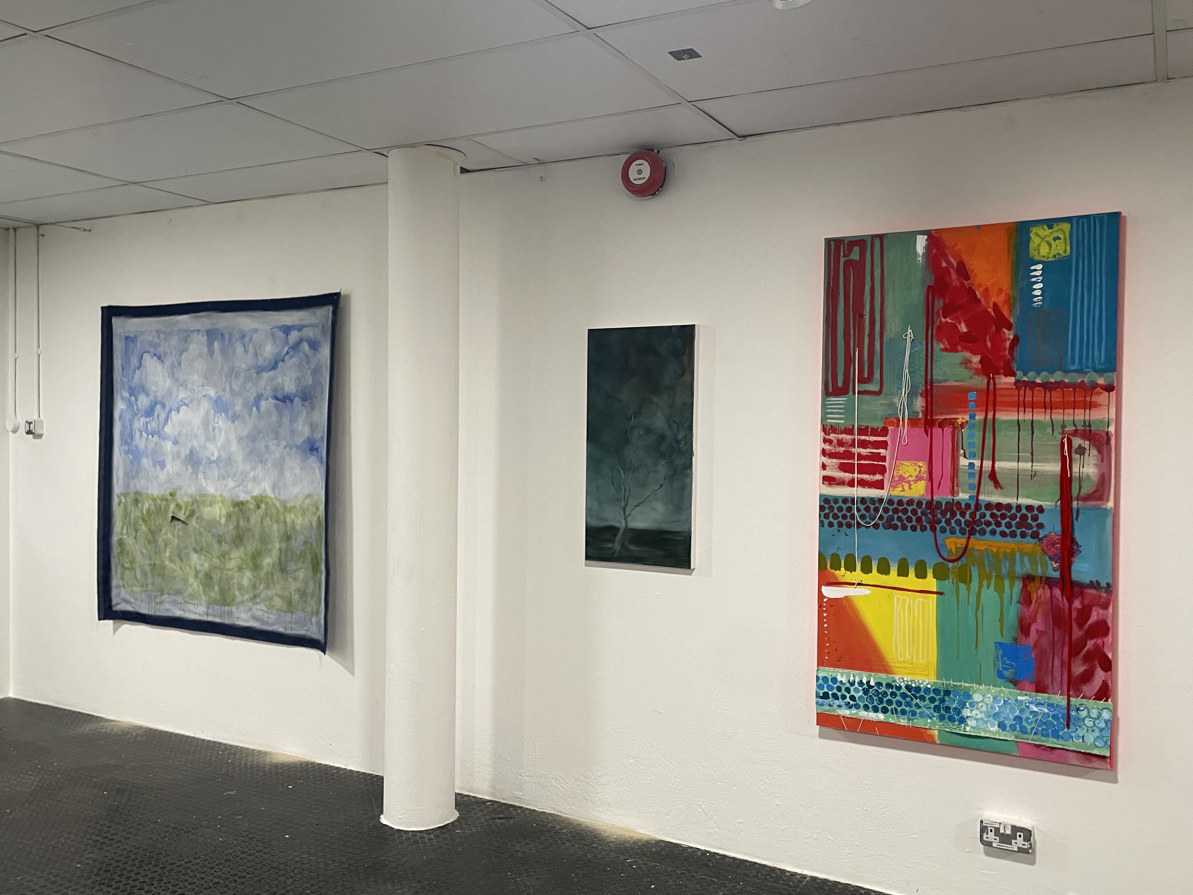 A gallery wall displaying three paintings: one with a cloudy sky and green field, one dark-toned abstract piece, and one colorful geometric artwork with bold patterns and drips.