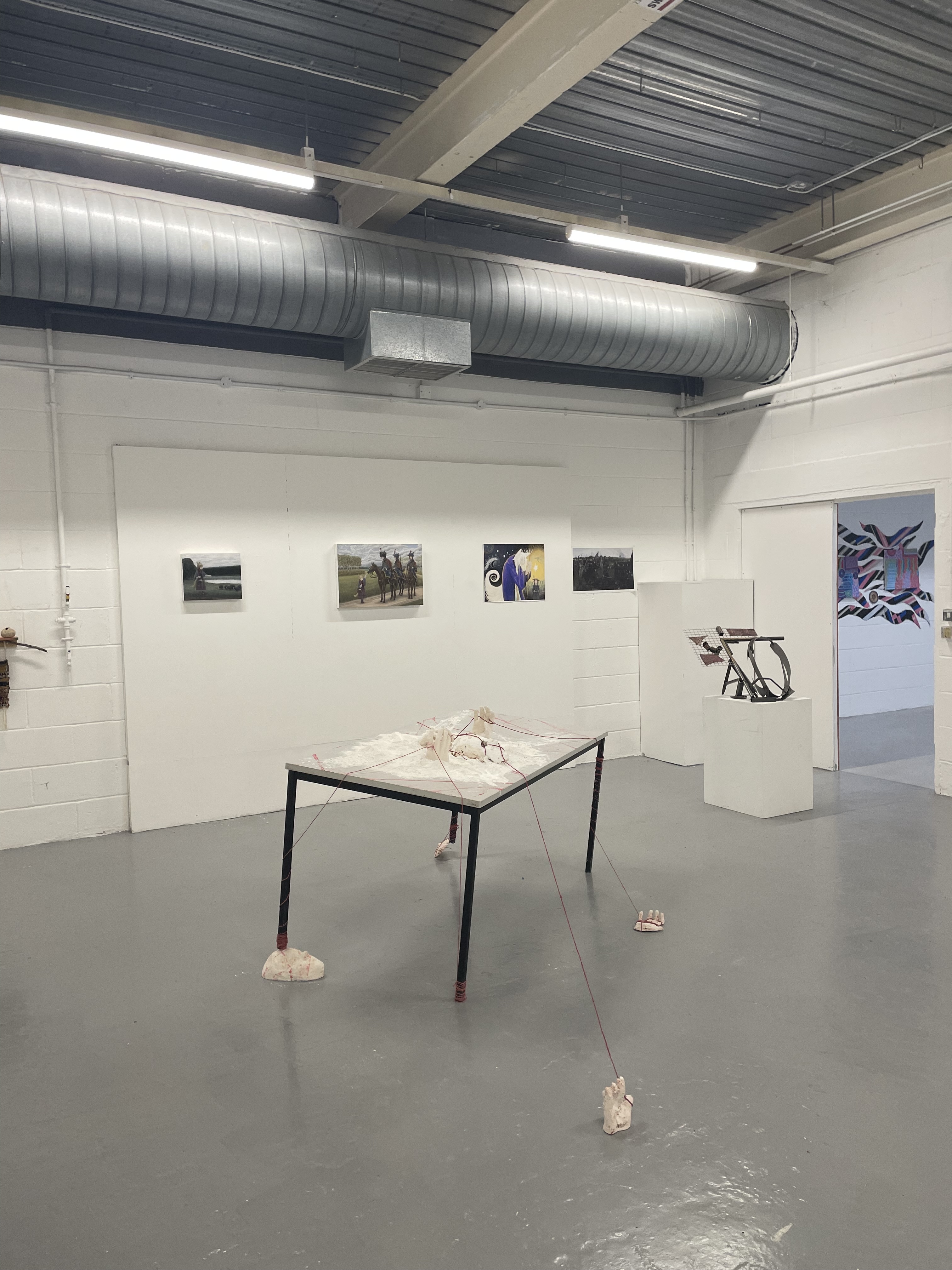 A contemporary art gallery with white walls and a gray floor, featuring a central table installation with objects tied by red strings and several photographs and sculptures displayed on the walls and pedestals.