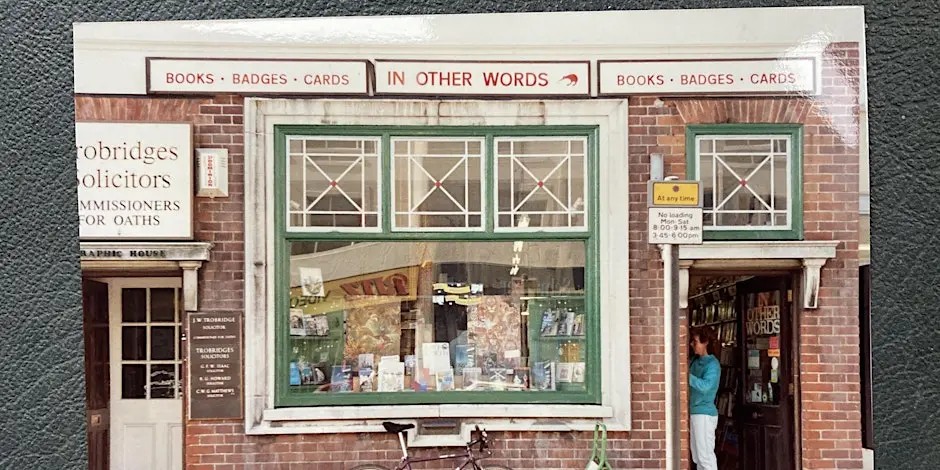  A storefront with a large green-framed window and two doors, displaying books inside. The sign above reads “IN OTHER WORDS” with “BOOKS · BADGES · CARDS” on both sides, and a bicycle is parked in front of the shop.