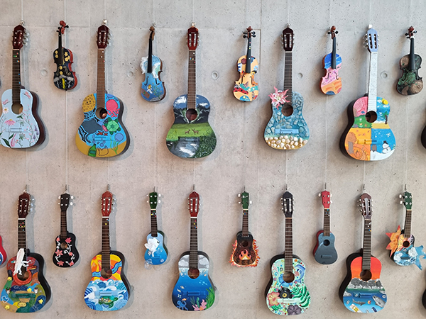 This image shows a display of repurposed acoustic guitars and violins mounted on a concrete wall, each transformed into a unique piece of artwork. The instruments have been painted by different artists or groups, featuring a variety of colourful designs including landscapes, abstract patterns, flowers, wildlife, underwater scenes and geometric motifs. Arranged in neat rows, the exhibition highlights creativity, sustainability and the reuse of musical instruments that would otherwise have been discarded. The neutral concrete background draws attention to the vibrant colours and individuality of each decorated instrument.