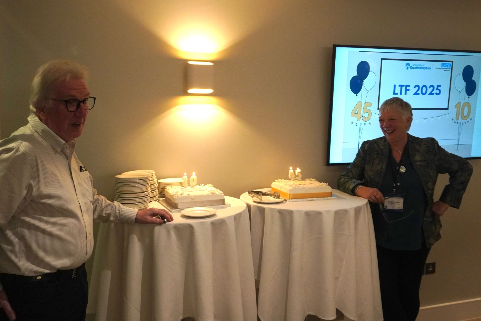 Two people stand beside tables holding the celebration cakes, with a screen in the background displaying “LTF 2025” and the numbers 45 and 10. Stacks of plates and lit candles are also visible on the tables.