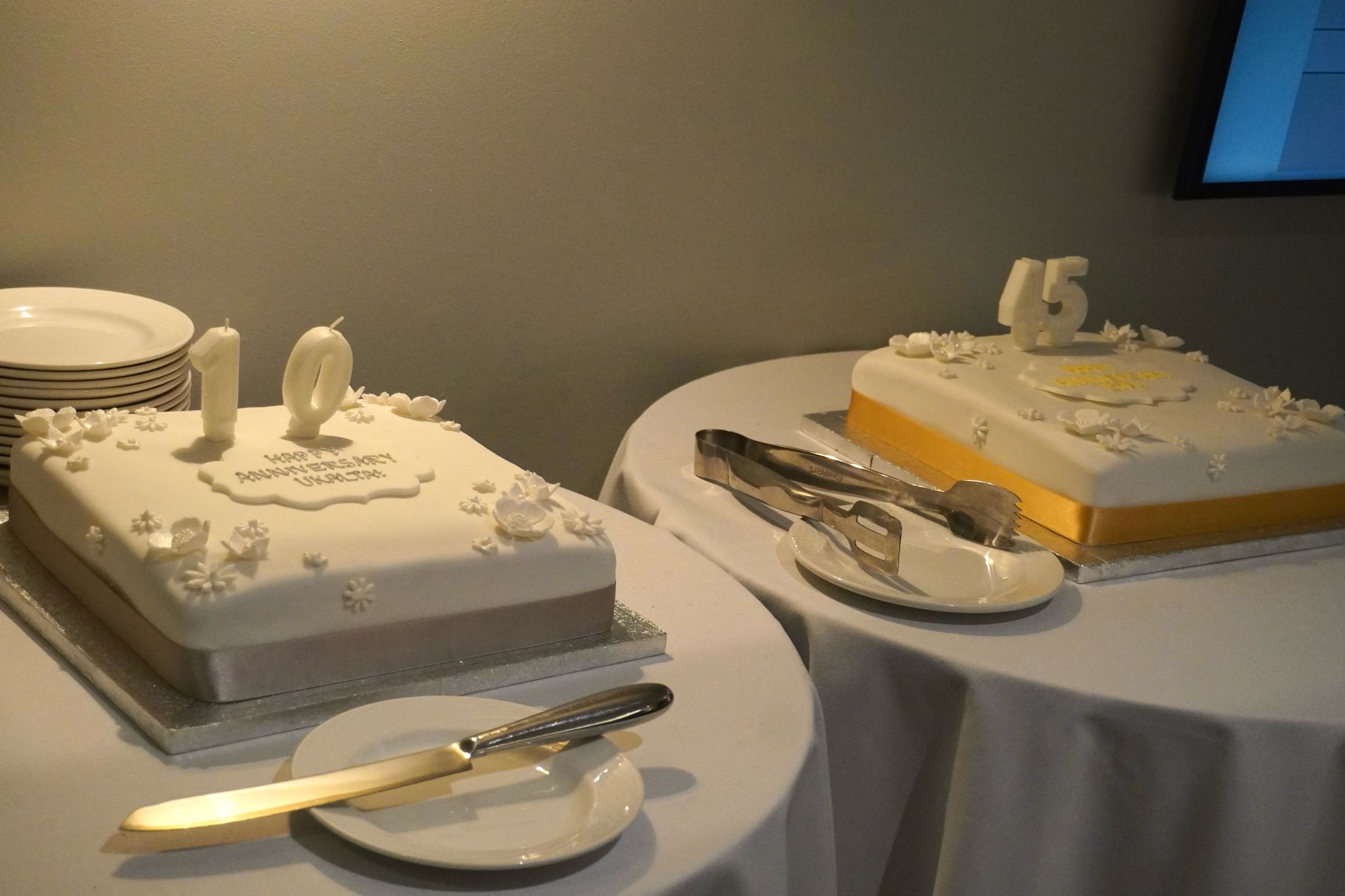  Two square celebration cakes with white icing and decorative flowers are displayed on round tables, one marked with the number 10 and the other with 45. Plates, a knife, and serving tongs are placed nearby.