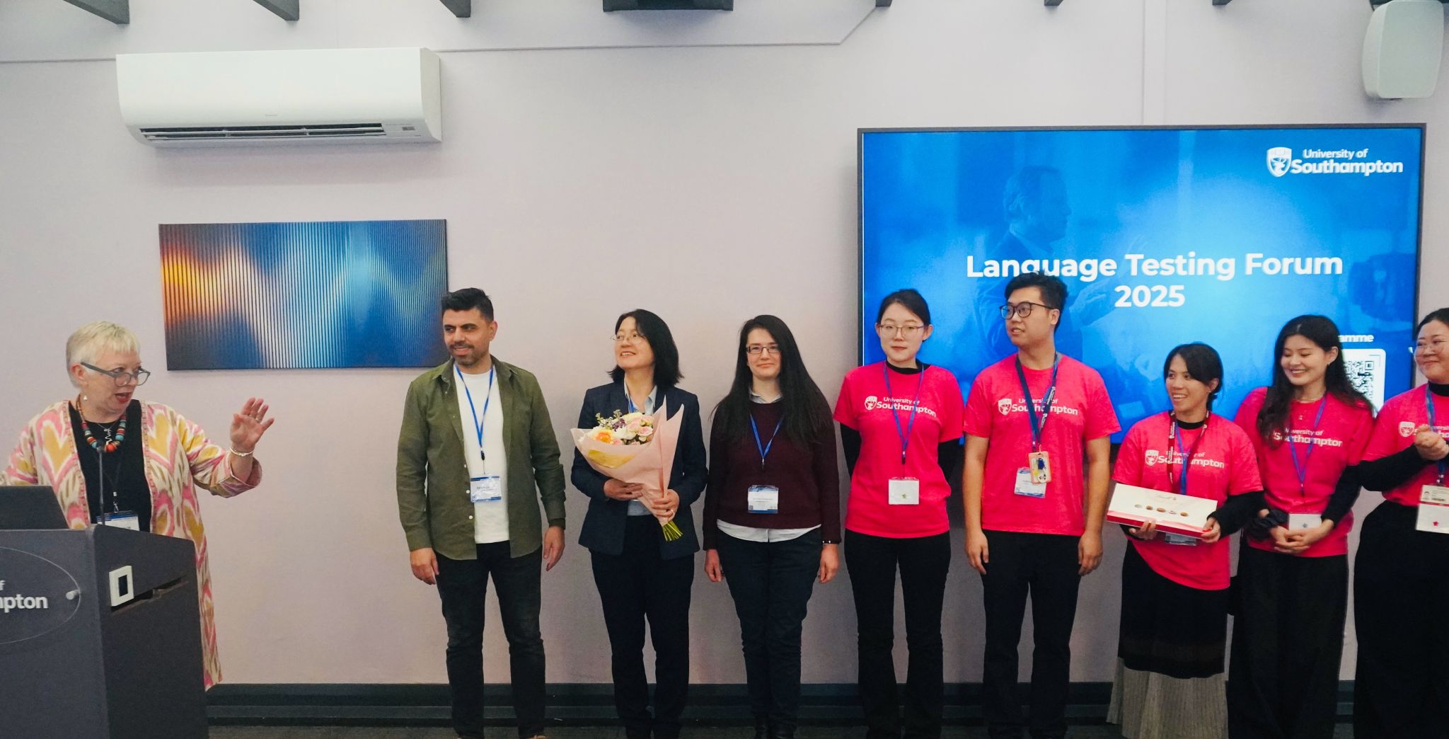 A group of people stands at the front of a room during the Language Testing Forum 2025, with one person holding a bouquet and others wearing bright pink T-shirts. A large screen behind them displays the event name.