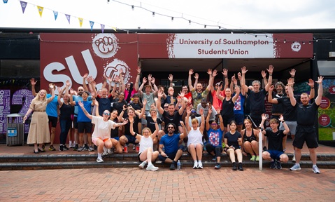 A large group of people wearing sports kit gather outside the University of Southampton Students' Union building, posing together with raised arms
