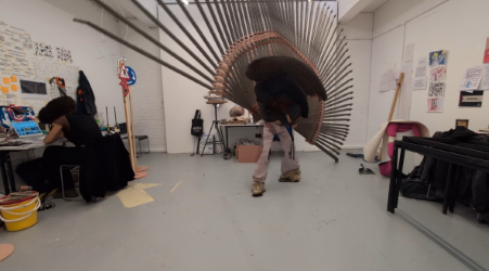 In a studio workspace, one person bends forward carrying a large sculptural structure made of radiating rods forming a circular, fan‑like shape. Another person sits at a desk nearby working on a computer, with tools, posters and materials scattered around the room.