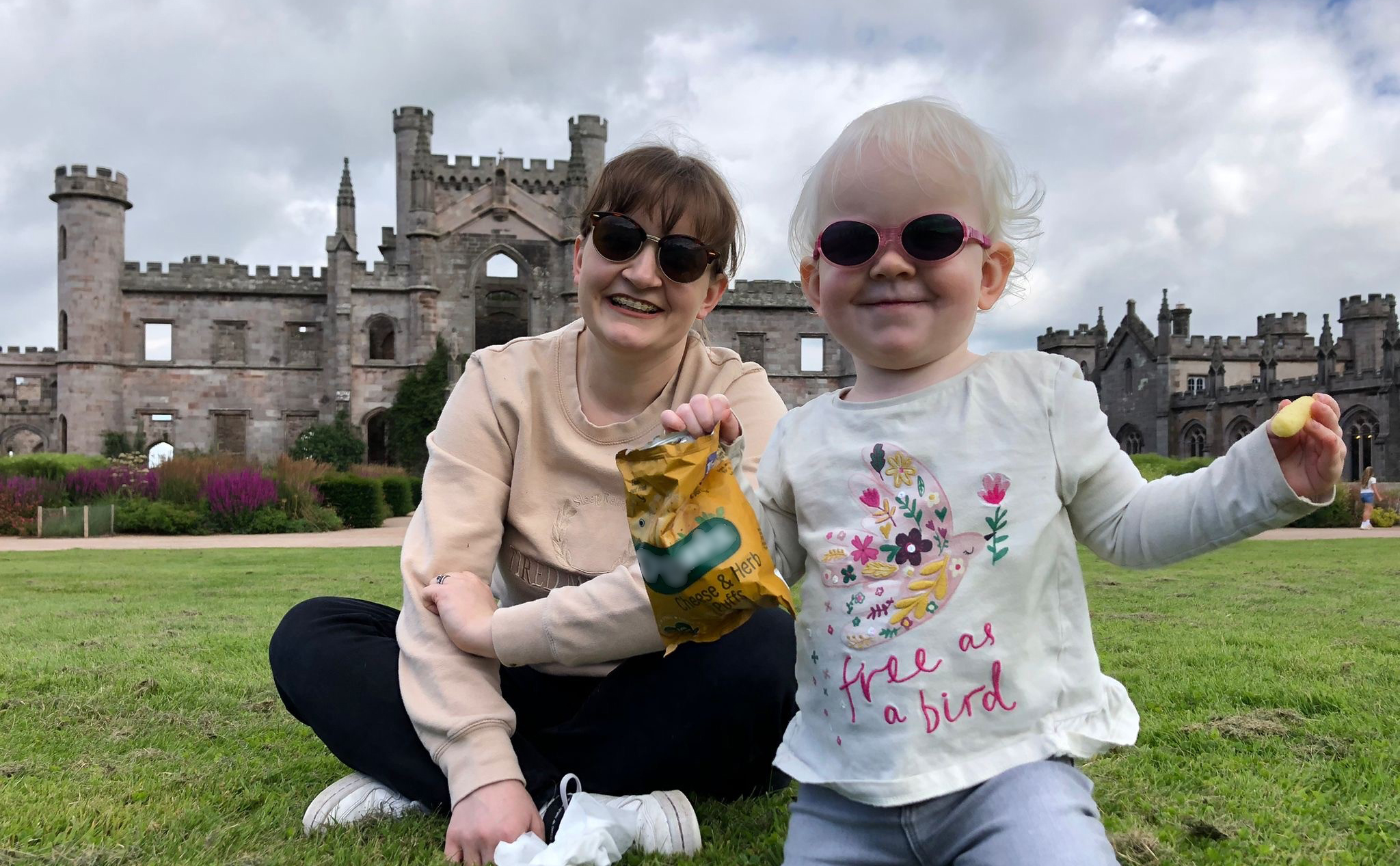 A young girl wearing dark glasses is kneeling on a lawn in front of her Mum who is sitting cross-legged with a castle in the background.