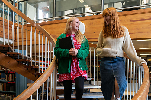 Two female students walk down a spiral staircase in a library. The students are smiling at one another and the one on the left in a green jumper is holding a book.
