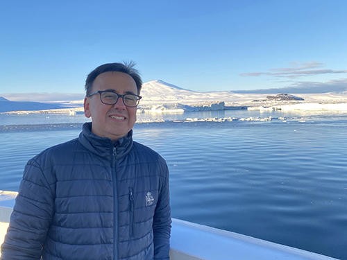 A man wearing glasses and a dark padded jacket stands on a boat in a frozen landscape, with calm blue water, snow-covered mountains and icebergs under a clear blue sky.