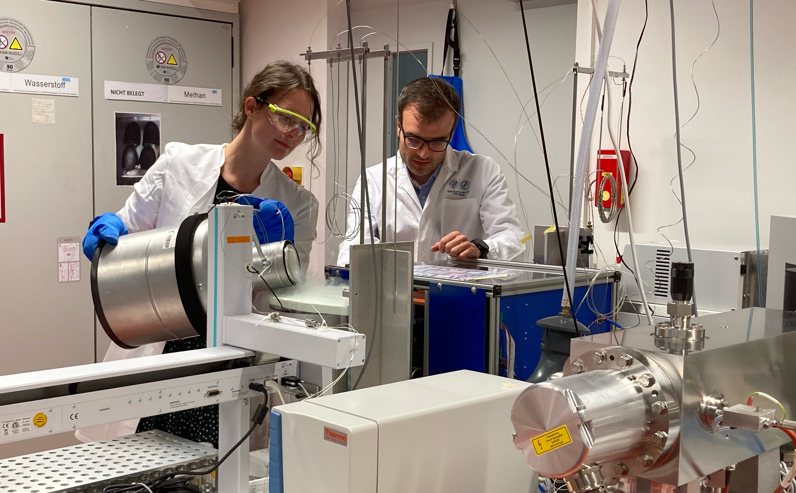 Scientists, a woman and man, wearing white coats in a lab, surrounded by technical equipment.