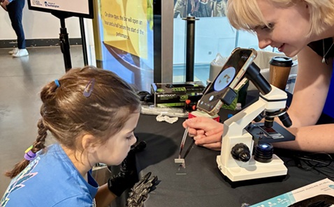 A young girl looking at a sample next to a microscope