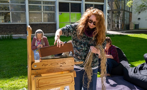 Woman with long hair wearing sunglasses, feeding straw-like stems into a wooden contraption and crushing them