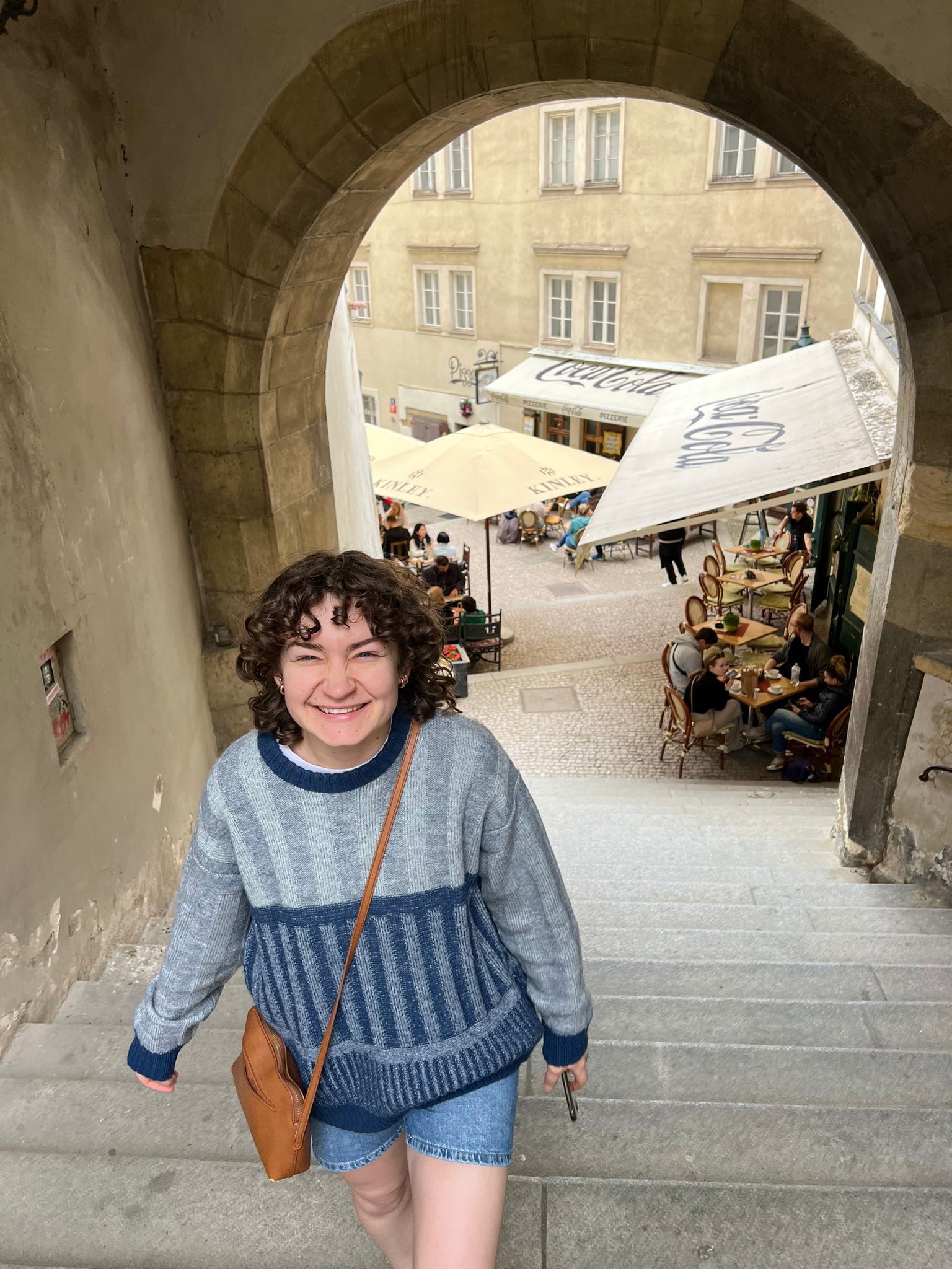 A person walks up a wide stone staircase beneath a large arched passageway overlooking an outdoor café courtyard. Historic buildings, café seating, and umbrellas are visible in the background.