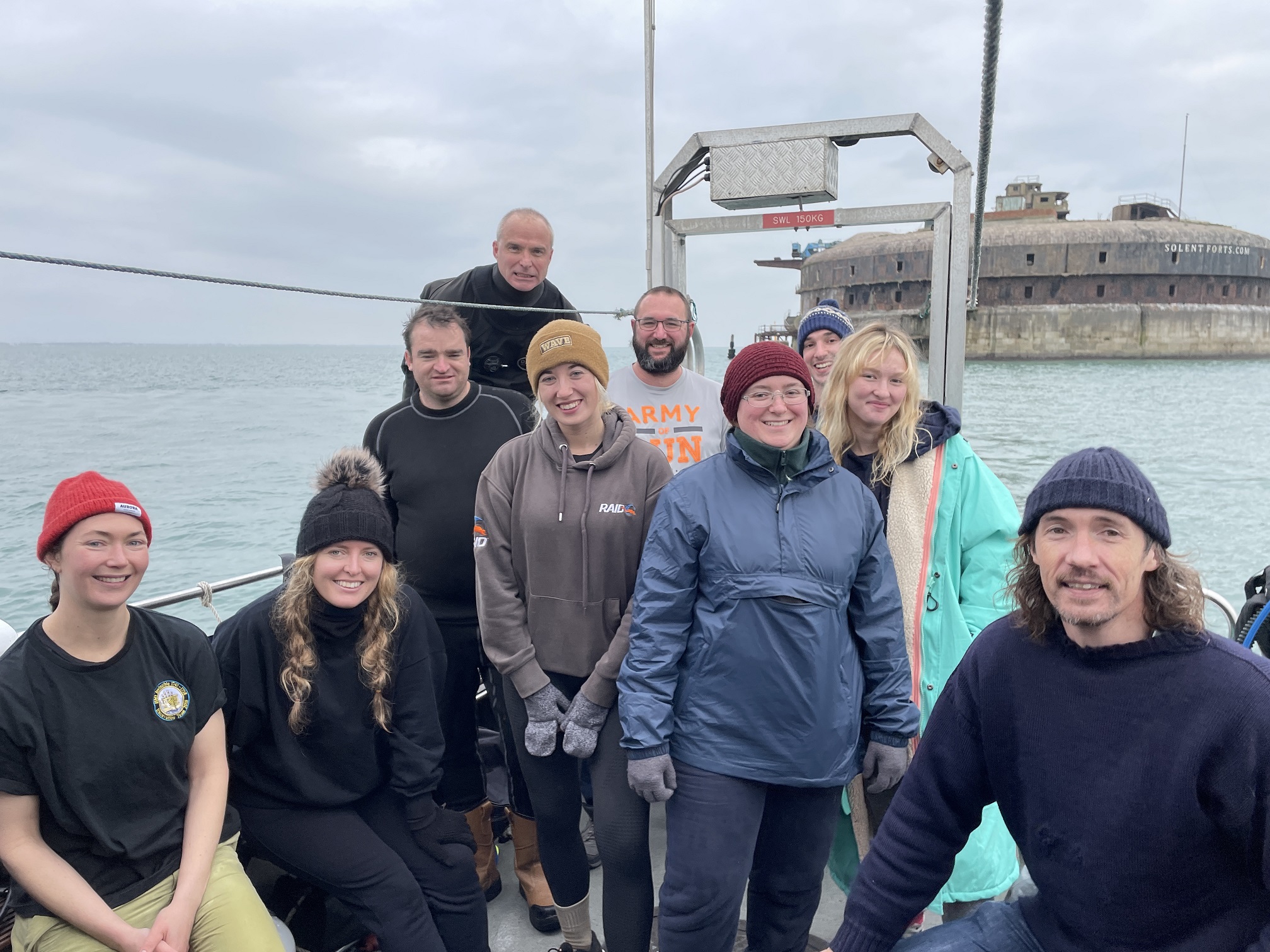 A group of ten people is posing for a photo on a boat in front of a historic maritime fort. The weather appears cold and overcast, with grey skies and calm waters. The group is dressed in warm clothing, including hats, gloves, and jackets, and most are smiling at the camera. 