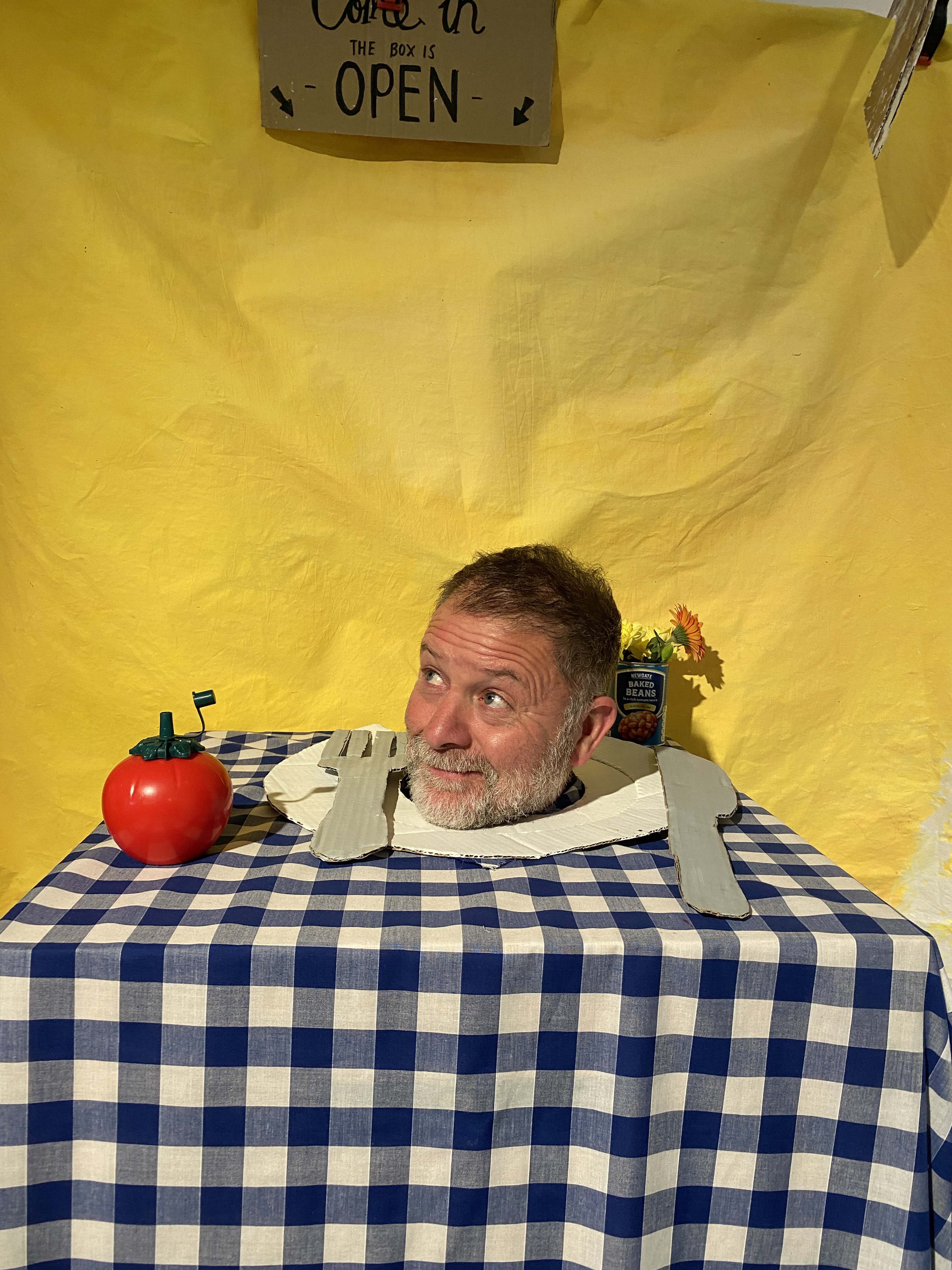 An art installation featuring a table covered with a blue and white check table cloth. There is a plate and tomato-shaped ketchup bottle, and an over-sized knife and fork. A man is standing under the table, with only his head visible on the plate. 