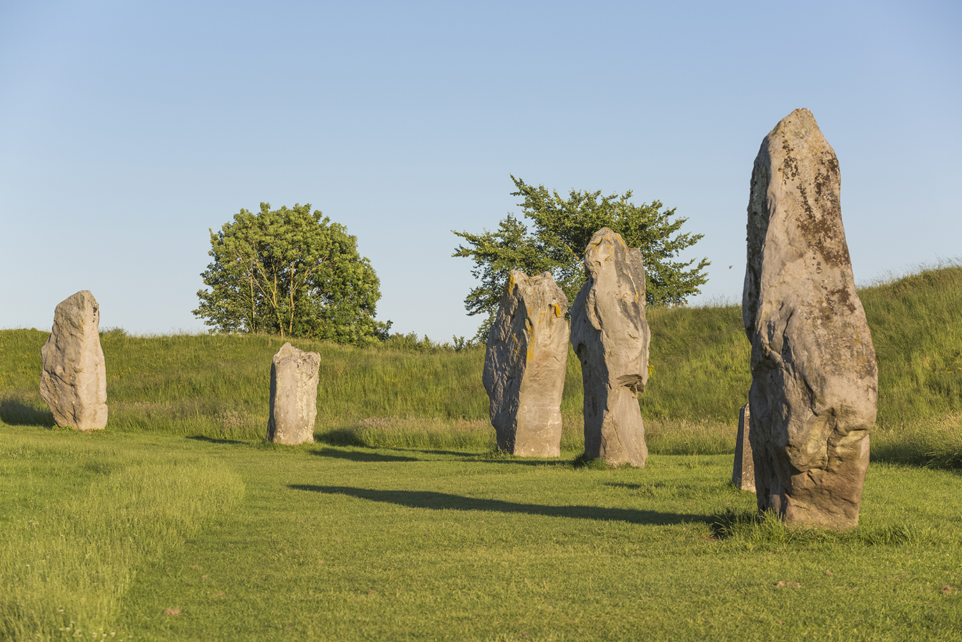 A section of the Avebury stone circle in Wiltshire, England, photographed on a sunny day. Several large, weathered standing stones are arranged in a loose arc on a grassy field. The stones cast long shadows in the afternoon light. Behind them, green grassy banks and a few leafy trees stand against a clear blue sky.