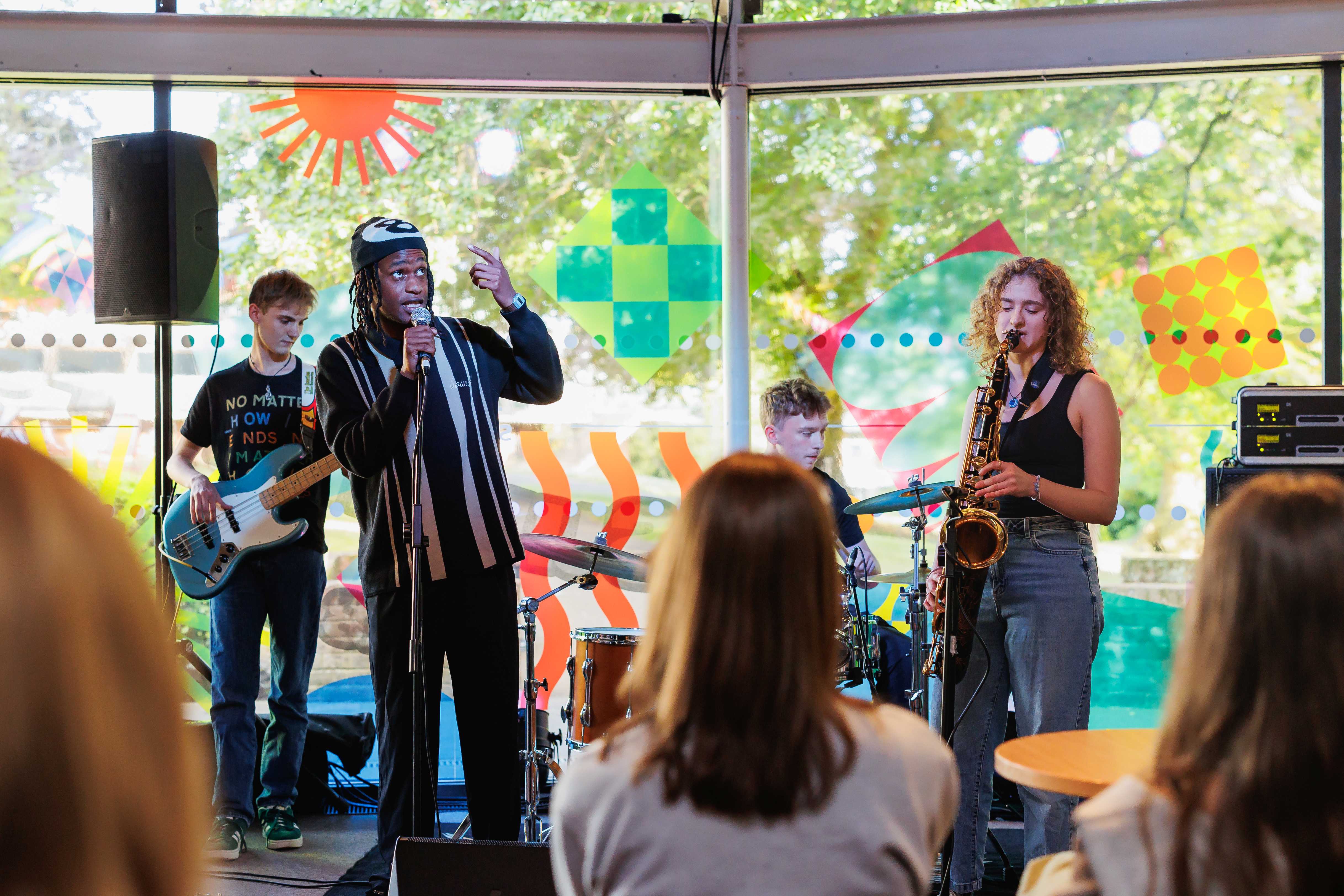 A small band performs on an indoor stage in front of a seated audience, with a vocalist at a microphone, a saxophonist, a guitarist, and a drummer. Bright, colourful geometric window decals and trees visible outside frame the performance space