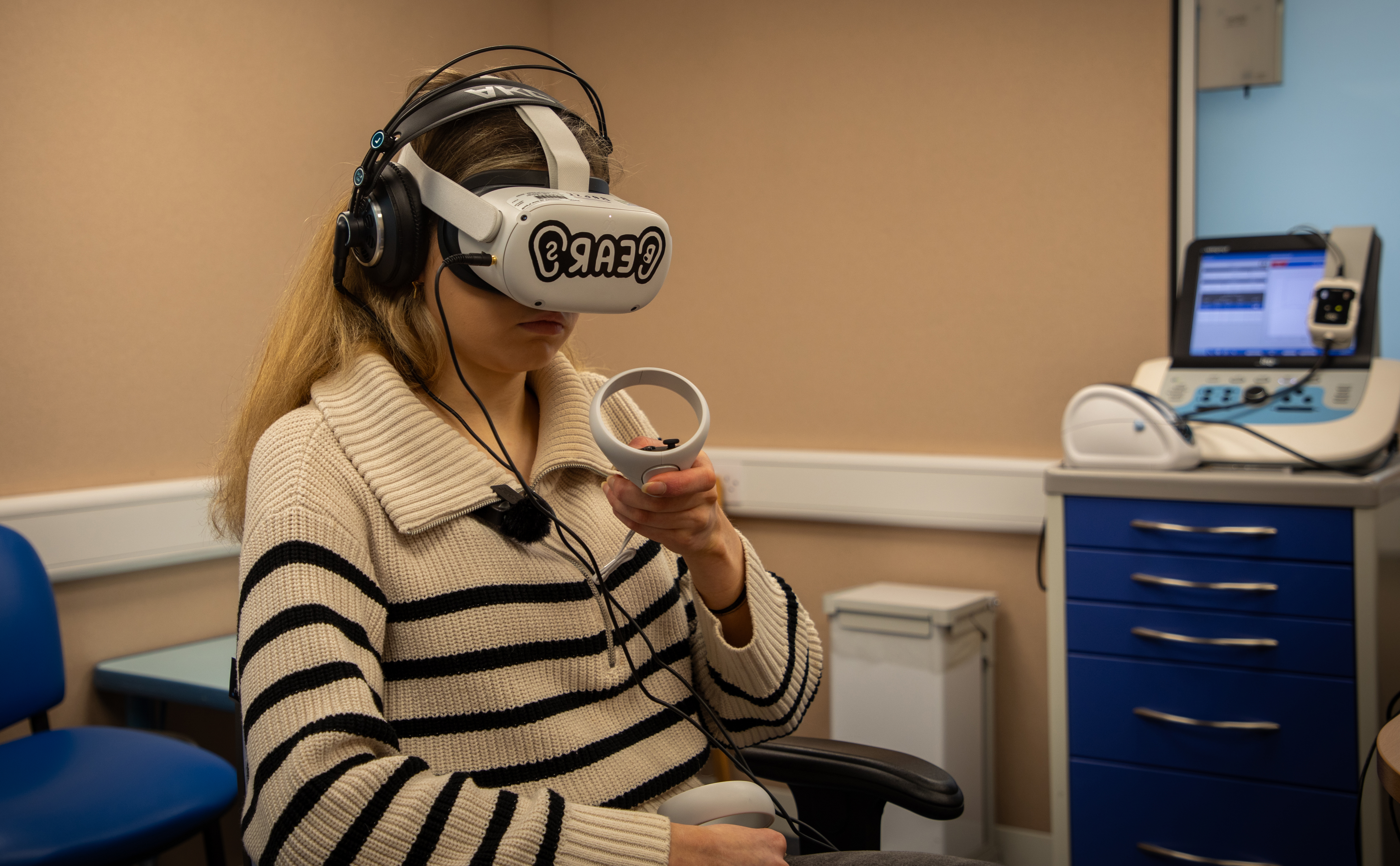 A woman sat in a chair with a virtual reality headset on, holding a VR control stick in her right hand.