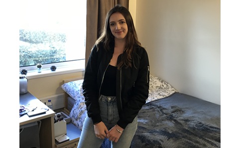 Young woman with long dark hair standing in front of a bed, next to a large window
