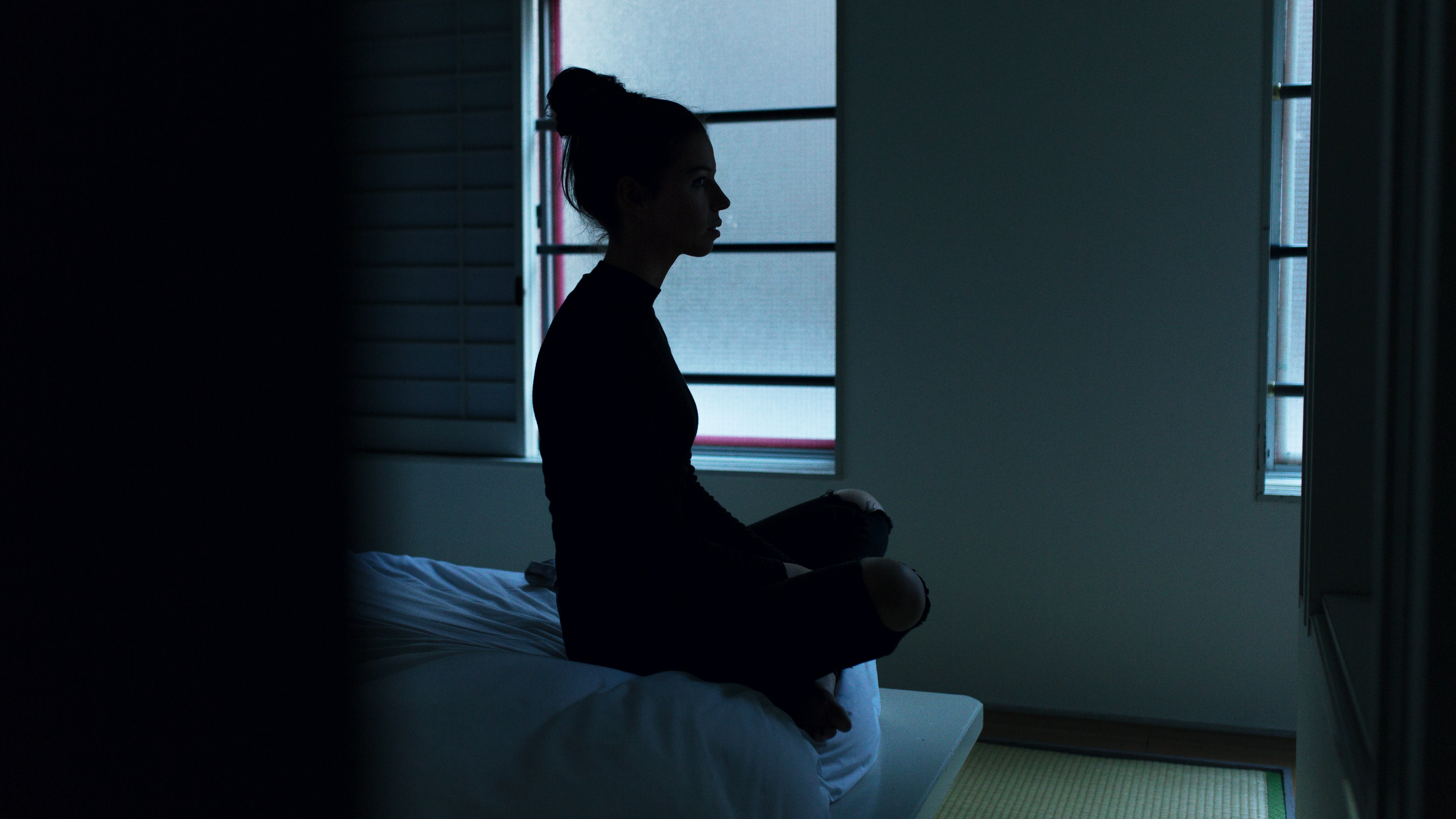 A woman dressed in black sits cross-legged on the edge of a bed in a dimly lit room, facing a window. Her profile is silhouetted against the soft light coming through the frosted glass.